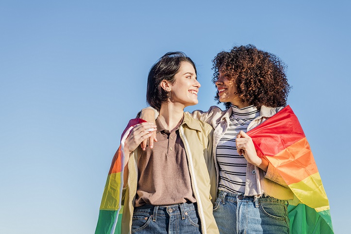  Duas mulheres se olhando e sorrindo enquanto seguram a bandeira do orgulho LGBTQIA+